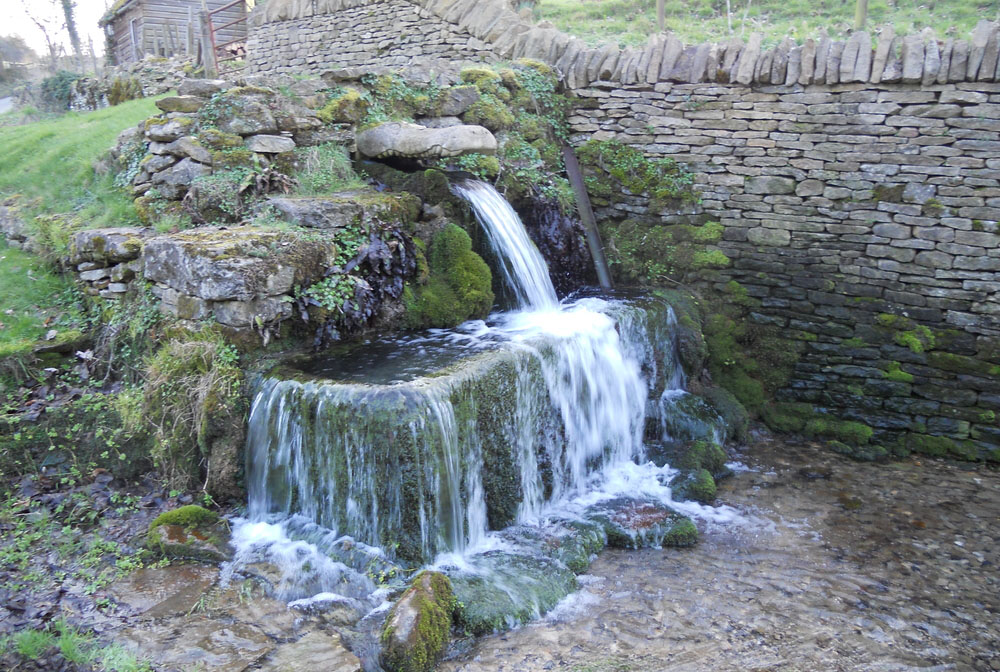 Water pours from a stone-built spring next to a drystone wall. The source of the water is a stone carved into the shape of a crocodile head and the water is coming out of its mouth.