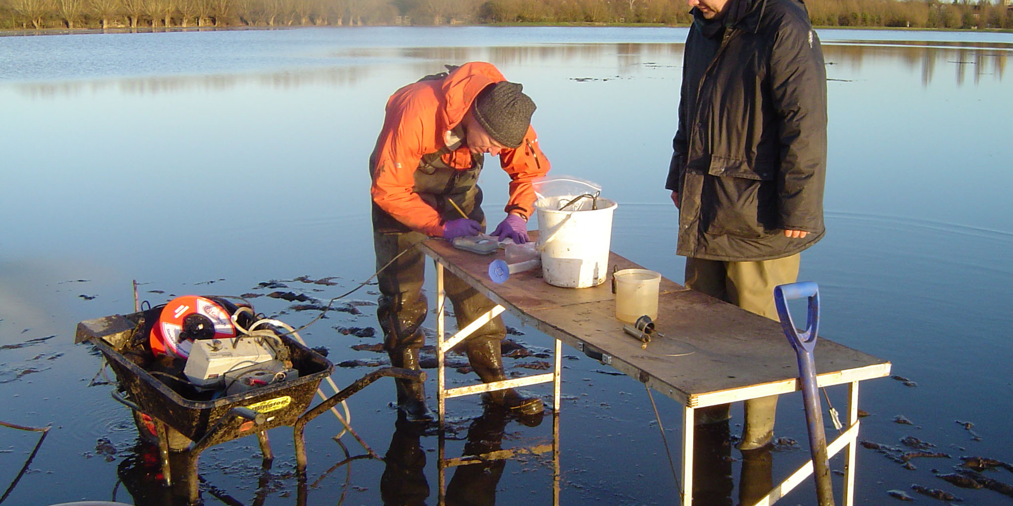Two white men stand next to a table and a wheelbarrow in a flooded field. There are buckets, tubes, cables and measuring equipment in the wheelbarrow and on the table.