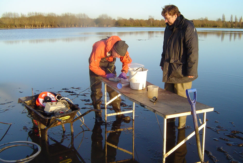 Two white men stand next to a table and a wheelbarrow in a flooded field. One man is wearing an orange waterproof coat and is bending over, writing in a notebook, while the other man, who is wearing black waterproof clothes, watches. There are buckets, tubes, cables and measuring equipment in the wheelbarrow and on the table.