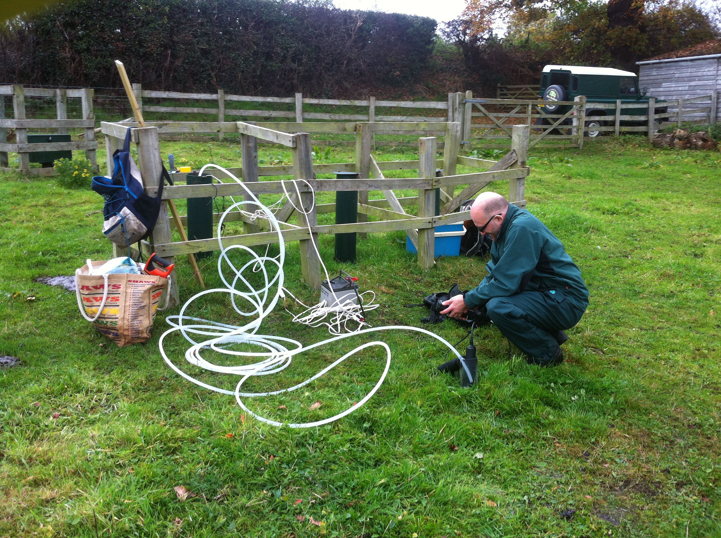 A white man wearing dark green clothes crouches next to a fence-off area in a field. There are tubes, bags, buckets and scientific equipment around him. He is looking at a hand-held instrument.
