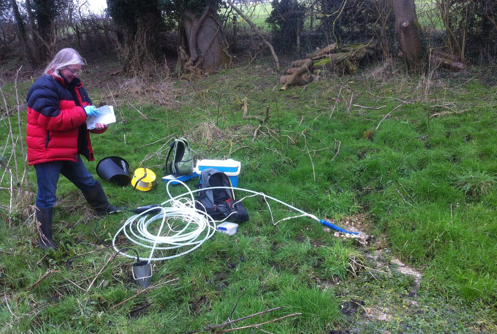 A white woman in a red and black coat is standing next to a spring writing in a notebook. A coiled tube leads from the stream to some water sampling equipment.