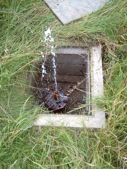 Water spurts from a borehole set in a rectangular concrete hole in some grass. rectangular