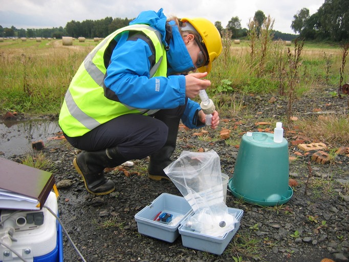 A white woman wearing a yellow hard hat, a blue jacket and a yellow hi-vis vest pours water into a bottle. She is crouched down and surrounded by buckets and sampling equipment.