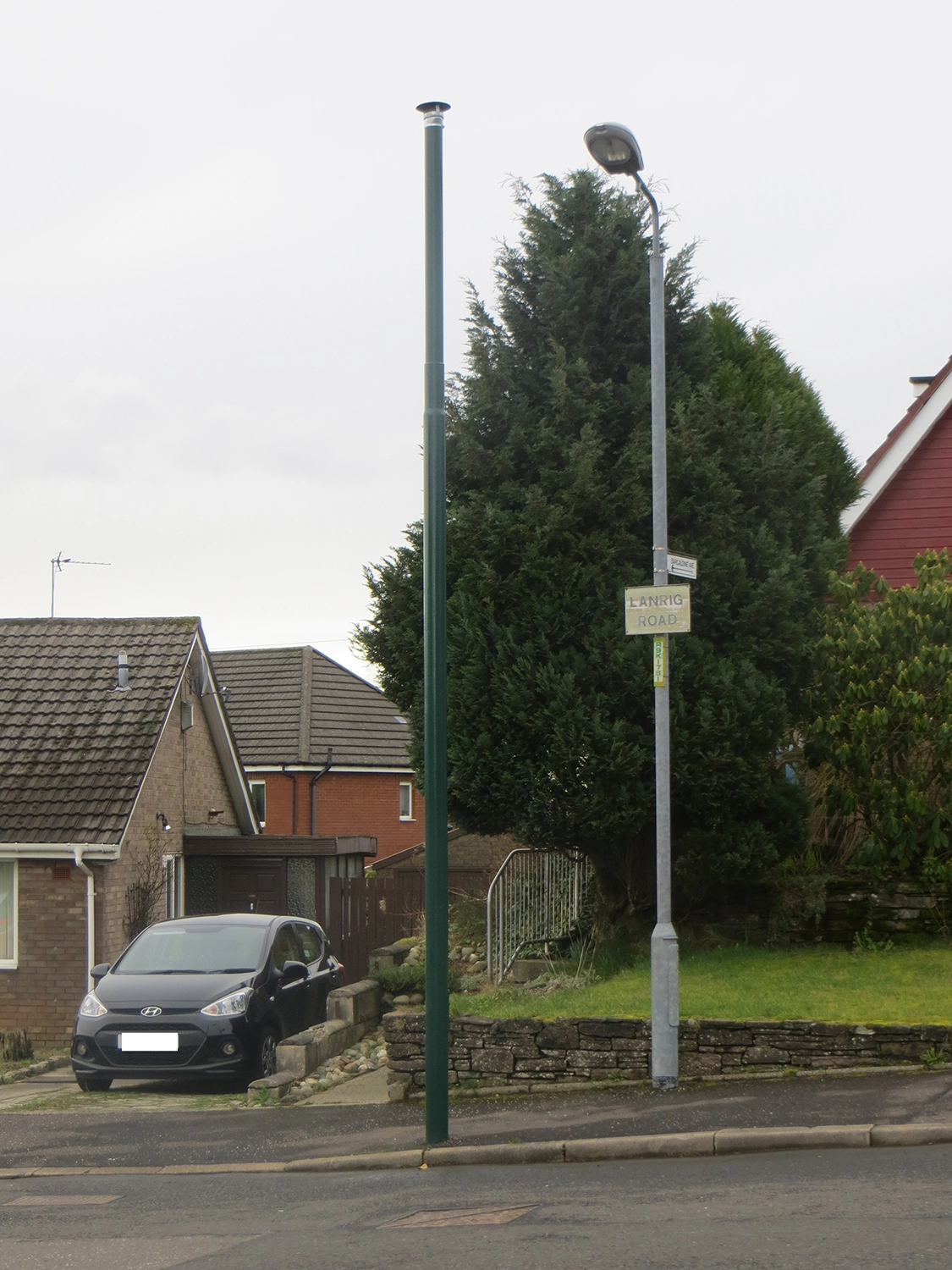 Looking towards a house with a car on the driveway and a large tree in the garden. There is a lamp post and a tall, green cylinder that is venting gas out of the ground.