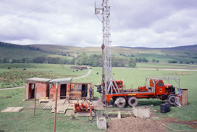 A tall drill rig on a red truck next to a log cabin. There are two white men in hard hats and overalls looking at the rig. The picture was taken in 1973.