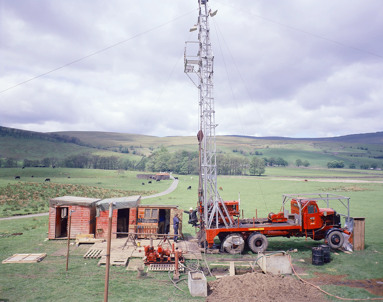 A tall drill rig on a red truck next to a log cabin. There are two white men in hard hats and overalls looking at the rig. The picture was taken in 1973.