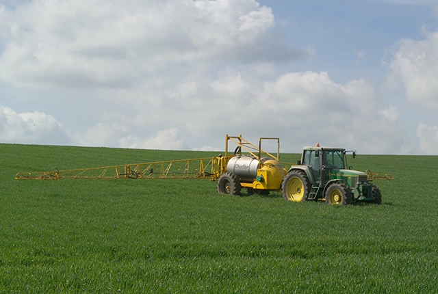 a yellow tractor pulls a tank behind it with crop spraying apparatus attached. It is moving across a green field.