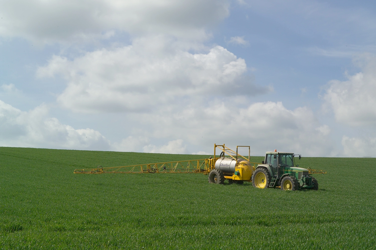 a yellow tractor pulls a tank behind it with crop spraying apparatus attached. It is moving across a green field.
