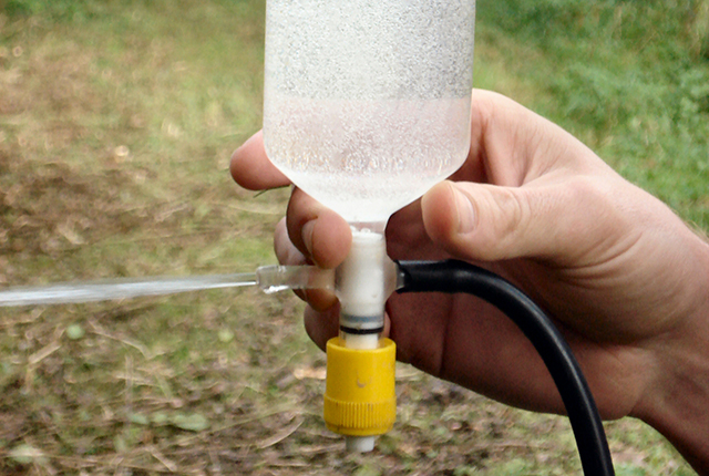 A white person's hand holding up a bottle containing a clear, colourless liquid with gas bubbling through it