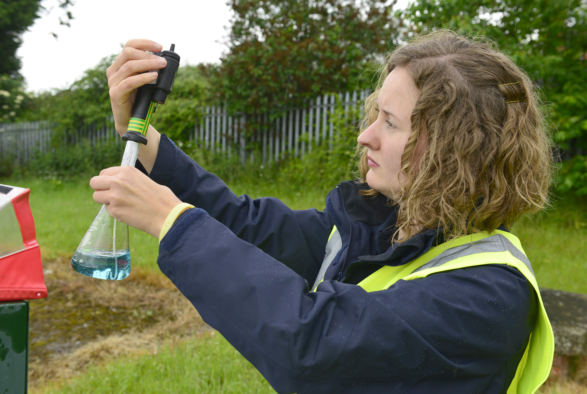A white woman wearing a dark blue jacket and a yellow hi vis vest holds up a conical flask which has a probe inserted into the blue liquid inside.