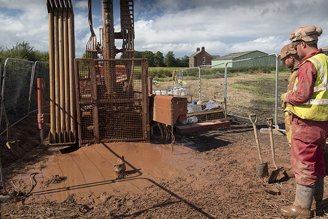 two men stand to the right watching a drilling rig in a very muddy field
