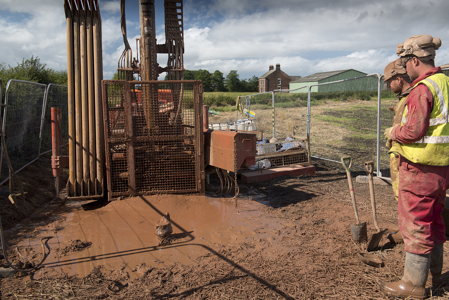 two men stand to the right watching a drilling rig in a very muddy field
