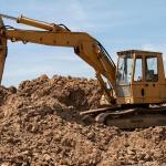 A large yellow digger excavating brown soil on top of a soil heap.