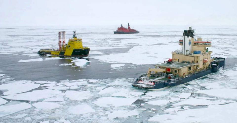 Three ships, one yellow, one red and one blue, float in an icy sea surrounded by fractured sea ice floes