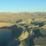 An aerial view of a sandy desert with a blue sky. There is a large depression filled with water, surrounded by roads across the sand and open-cast copper mines.