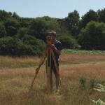 A man standing in a field of of yellow and green grass looking through a piece of equipment called a theodolite. The theodolite is standing on a yellow metal tripod. There are lush green trees and blue sky in the background.