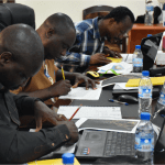 A group of students sat at computer terminals, drawing with pencils on pieces of paper