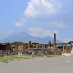 The wide, open forum of Pompeii, with ruined columns and tourists in the middle distance and the silhouette of the volcano Vesiuvius in the background.