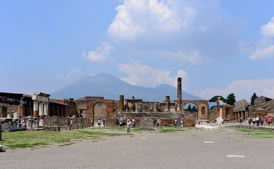 The wide, open forum of Pompeii, with ruined columns and tourists in the middle distance and the silhouette of the volcano Vesiuvius in the background.