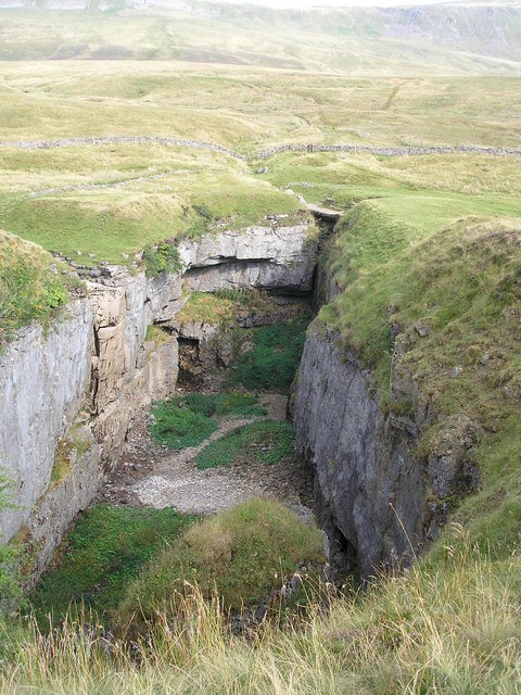 A large, rectangular depression in the ground with vertical walls of grey rock. There are green hills in the background and the floor of the depression and the land around it are covered in green grass.