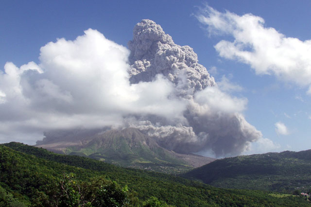 A dark coud of ash rises over the Sofrier Hill volvano in Montserrat. The volcano is obscured by white clouds and there are green hills in the foregrund.