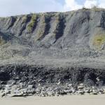 Gullies in a cliff face made of dark-coloured rock, forming a landslide on a beach