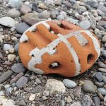 A large round pebble or orang brick with grey mortar running across it, sitting on a laer of smaller, grey, white and black pebbles and sand.