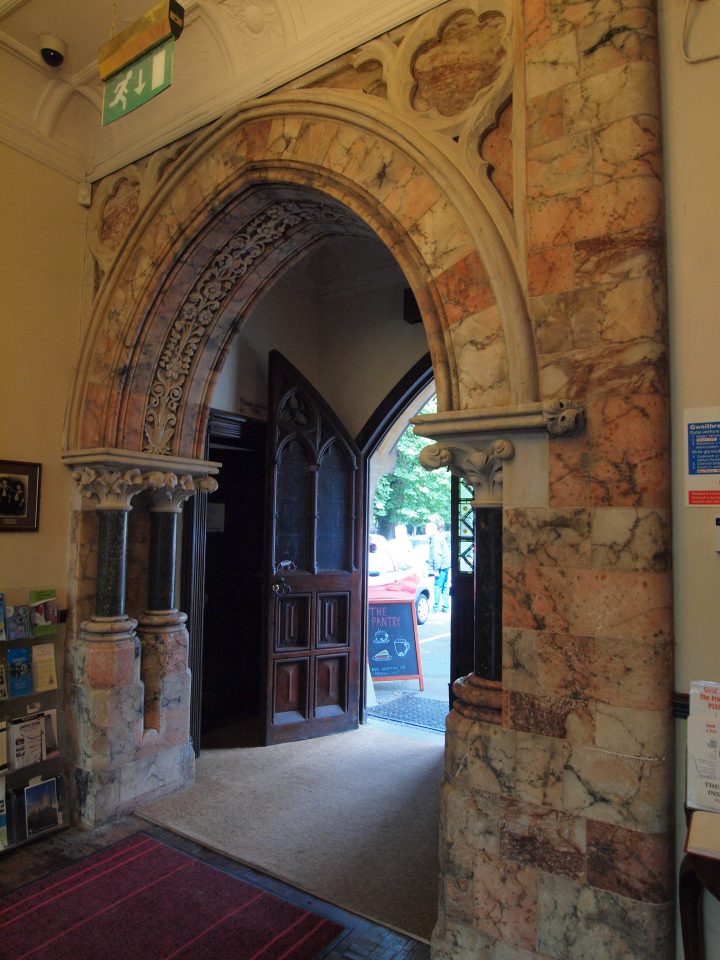 Arched doorway of pink and white alabster. There is a big wooden door leading outside.