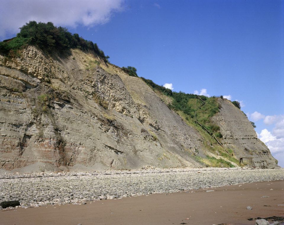 Cliffs of grey and greenish rcks with vegetation growing on them. At the foot is a pebbly beach grading down to sand in the foregrouns and a blue sky in the backgound.