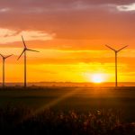 Tall wind turbines are silhouetted against an orange sky, with the sun setting on the horizon