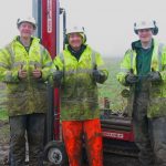 Three geologists wearing hi-vis gear and covered in mud, giving the camera a thumbs-up