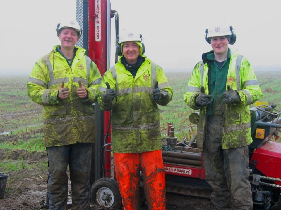 Three geologists wearing hi-vis gear and covered in mud, giving the camera a thumbs-up