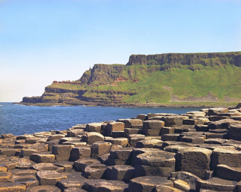 The Giant's Causeway in Northern Ireland. Hexagonal blocks of brownish grey rock in the foregroud, with seain the biddle ground and a high cliffs of basailt columns and green grass in the background.