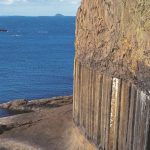 The Isle of Staffa, Scotland. A flat platform of brownish grey rock with the sea visible behind it, with straight vertical columns rising up out of it. The columns are overlain with more rock plaars that appear more twisted and convoluted.