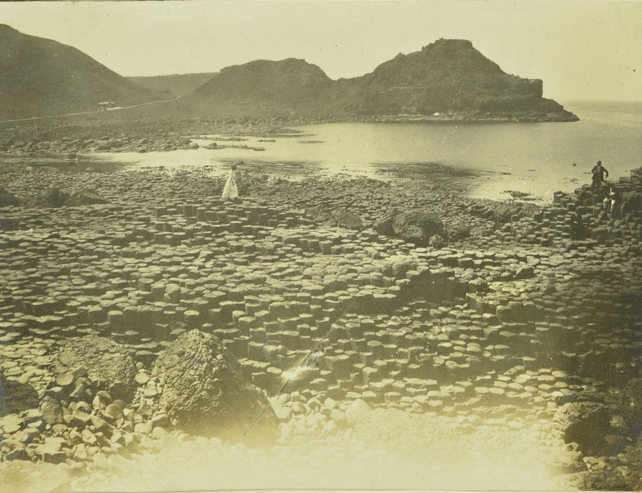 A black and white photograph from theearly twentiwth century showing a woman in a long white dressalking on the columns at the Giant's Causeway