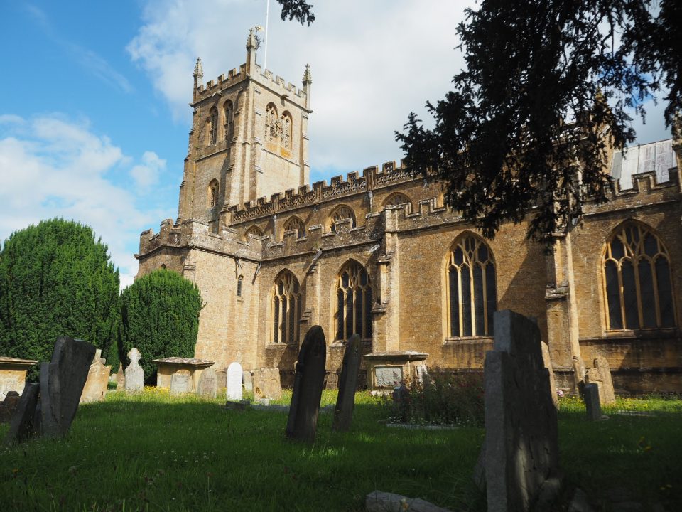 A large church made of light golden-coloured stone in a grassy graveyard with trees