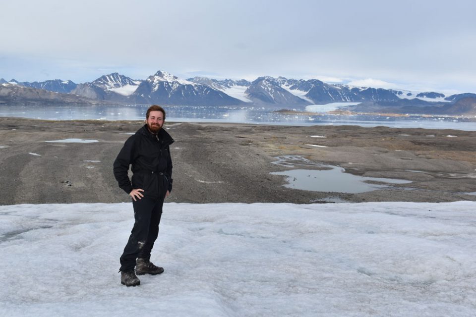 A white man with a beard dressed in black overalls standing on the ice of a glacier. There is a strip of bare rocks with a river behind him leading to the water of the fjord and snowy mountains in the distance.