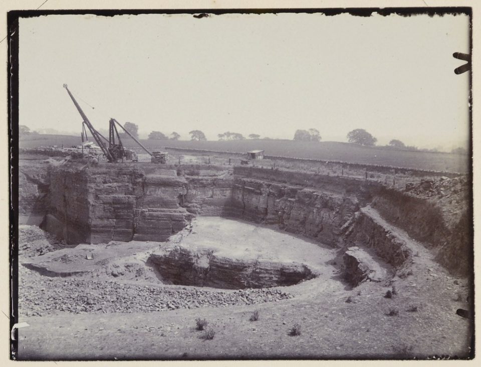 A black and white picture of a working quarry, showing the quarry faces and some machinery at the top of the working