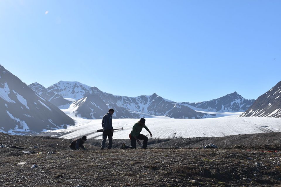 Three people, one crouching, one standing and one kneeling, on the rocky moraine in front of a white glacier. There are mountains in the distance with the glacier flowng between two of the peaks.