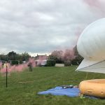 A white balloon and a white kite float above a yellow, ring-shaped inflatable, which is sitting on the ground on a blue tarpaulin. The kite and balloon are tethered to a pole. A person in a hi-vis vest stands to theleft holding a flare that is emitting pink smoke.