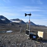 A wind turbine, a solar panel and a box cntaining sensor equipment installed on bare, rocky ground in front of snowy mountains.