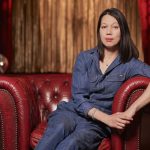 A woman with long dark hair dressed in a blue trouser suits sits in a red leather chair