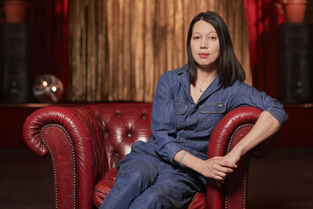 A woman with long dark hair dressed in a blue trouser suits sits in a red leather chair