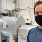 A young white man in a black face mask and abluer t shirt stands in front of scientific equipment consisting of a large barrel-shaped vessel surrounded by pipework