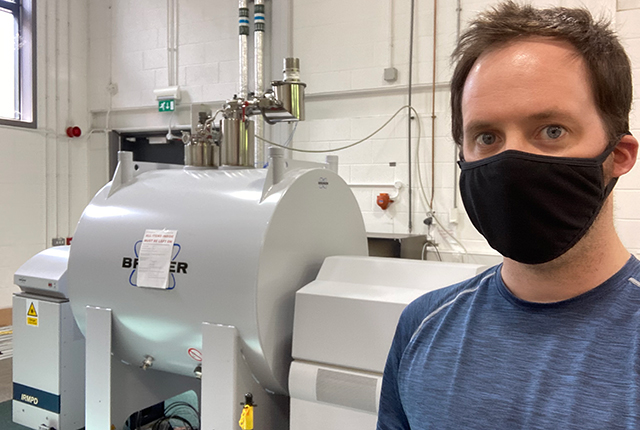 A young white man in a black face mask and abluer t shirt stands in front of scientific equipment consisting of a large barrel-shaped vessel surrounded by pipework