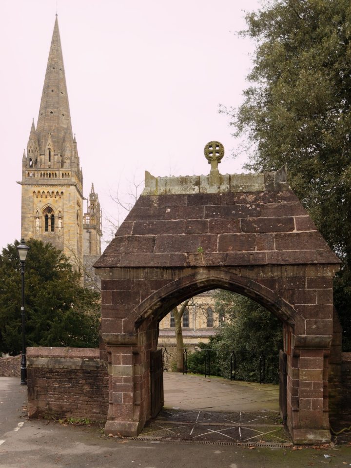 A tall spire of a cathedral rises in the backgroun. The roofed gate (lychgate) in the foreground