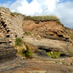 A snady and rocky cliff covered by a brick wall that is cru,mbling down onto a beach