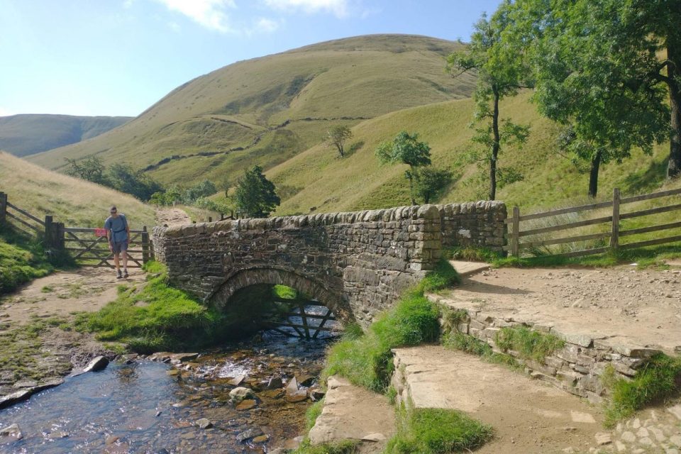 A narrow stone bridge with a single arch crossing a small river. A dome-shaped hill covered in grass rises in the background. There is a tree to the right of the bridge and a white person wearing shorts and a t-shirt to the left, standing in front of a wooden fence and gate.