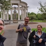 Three staff members in a park infront of a circular, colonnaded structed, holding litter-picking equipment