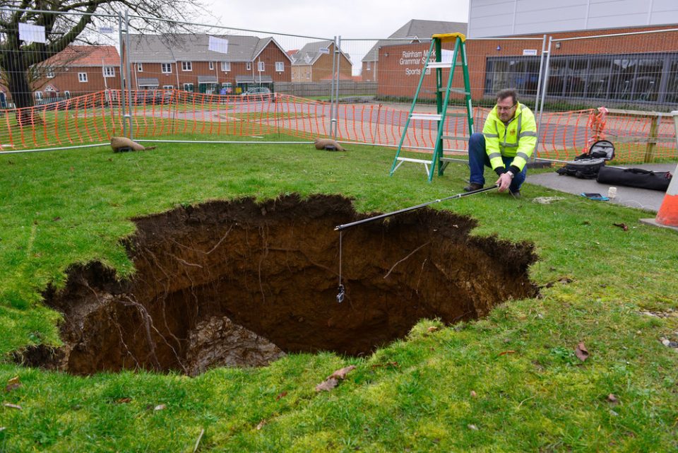 A deep hole in a a lawn, with a man in a hi-vis yellow vest kneeling next to it holding a measuring instrument over the hole